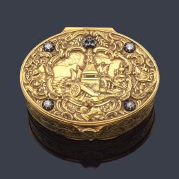 Lote 2593: Caja ovalada realizada en oro amarillo de 18K cincelado con motivo central escena de alegoría de la guerra con corona real y cartelas con trofeos, enriquecido con cuatro diamantes talla antigua.